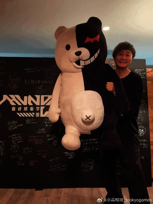 小高和刚与黑白熊玩偶合影，背景是电脑屏幕显示着游戏画面