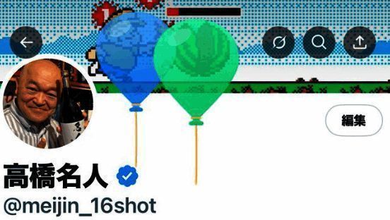 高桥名人近年工作照，手持游戏手柄，专注盯着电脑屏幕，身旁摆放多款游戏周边