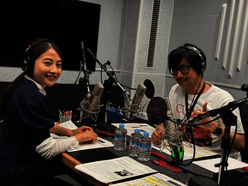 小岛秀夫在办公室内专注工作，身后是《死亡搁浅2》的游戏海报和开发设备