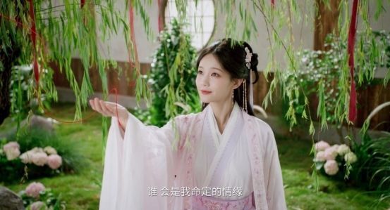 游戏中李一桐角色立绘，手持团扇微笑站立，身后是盛开的桃花树与木屋