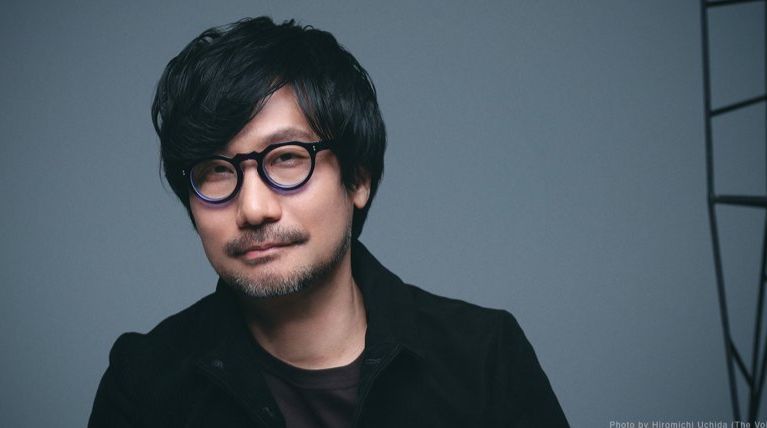 小岛秀夫在舞台上的经典形象，身穿标志性风衣，神情专注地望向远方