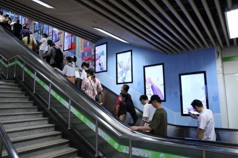 coser装扮成游戏角色在地铁站内与屏幕中的角色合影，周围乘客驻足拍照