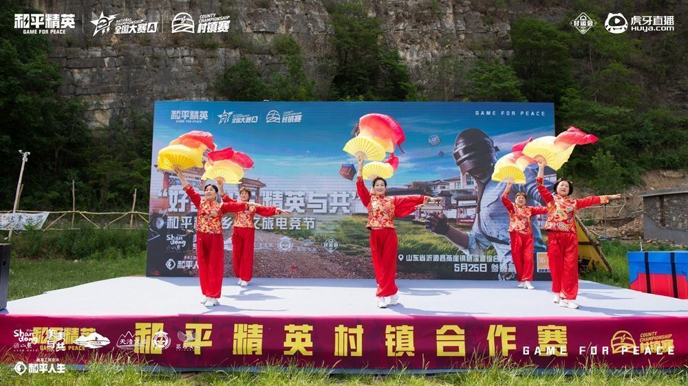 村民在和平精英比赛现场围观，大屏幕显示对战画面，背景有乡村舞台和横幅