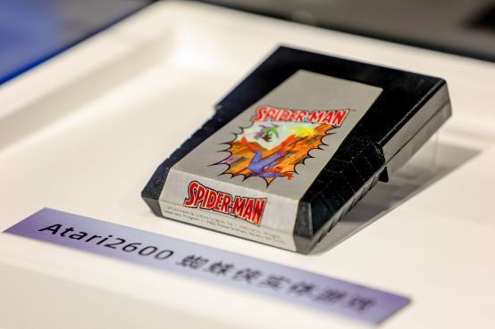 玻璃展柜中陈列着1982年雅达利2600平台的实体蜘蛛侠游戏卡带，旁边配有老式主机和电视模拟运行画面
