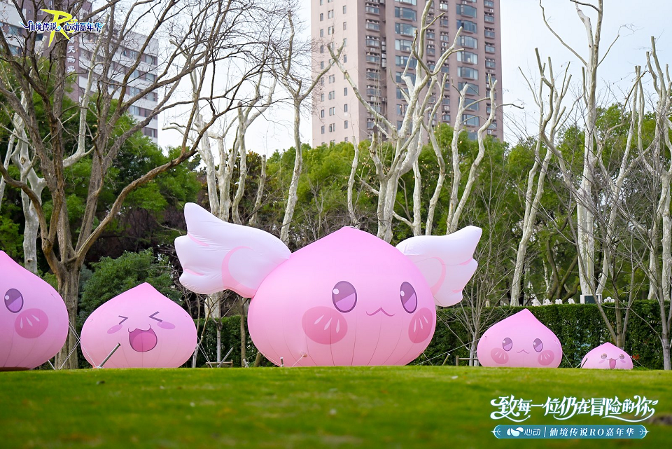 制作人邢万里手持手柄站在直播屏幕前，画面实时显示守爱2游戏内场景：角色在阳光草地上慵懒伸展，突然切换为猫形态翻滚撒娇，动作流畅萌感十足